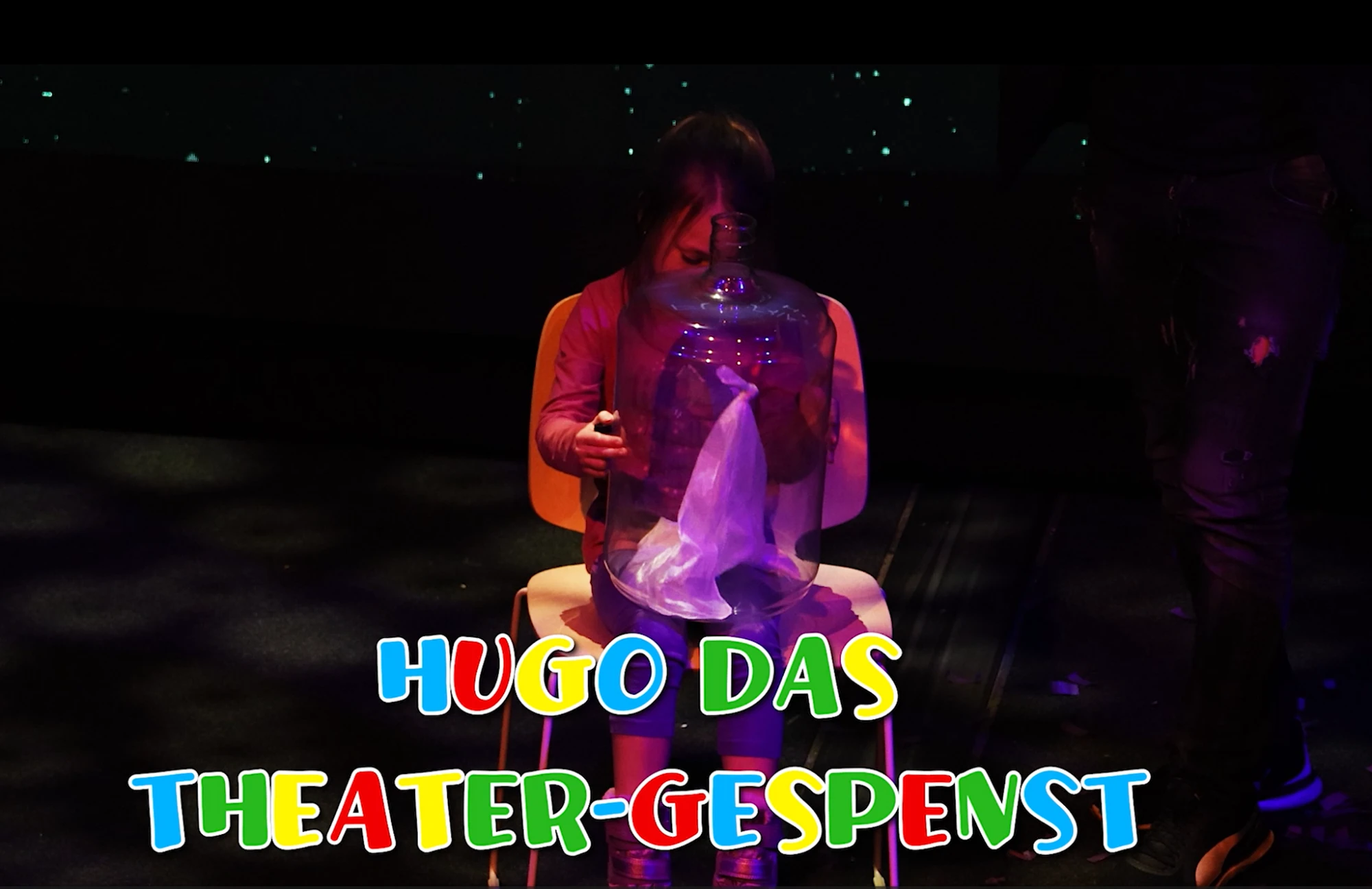 Ein Mädchen sitzt auf der Bühne vor einem Glas, in dem Hugo, das Theater-Gespenst, schwebt